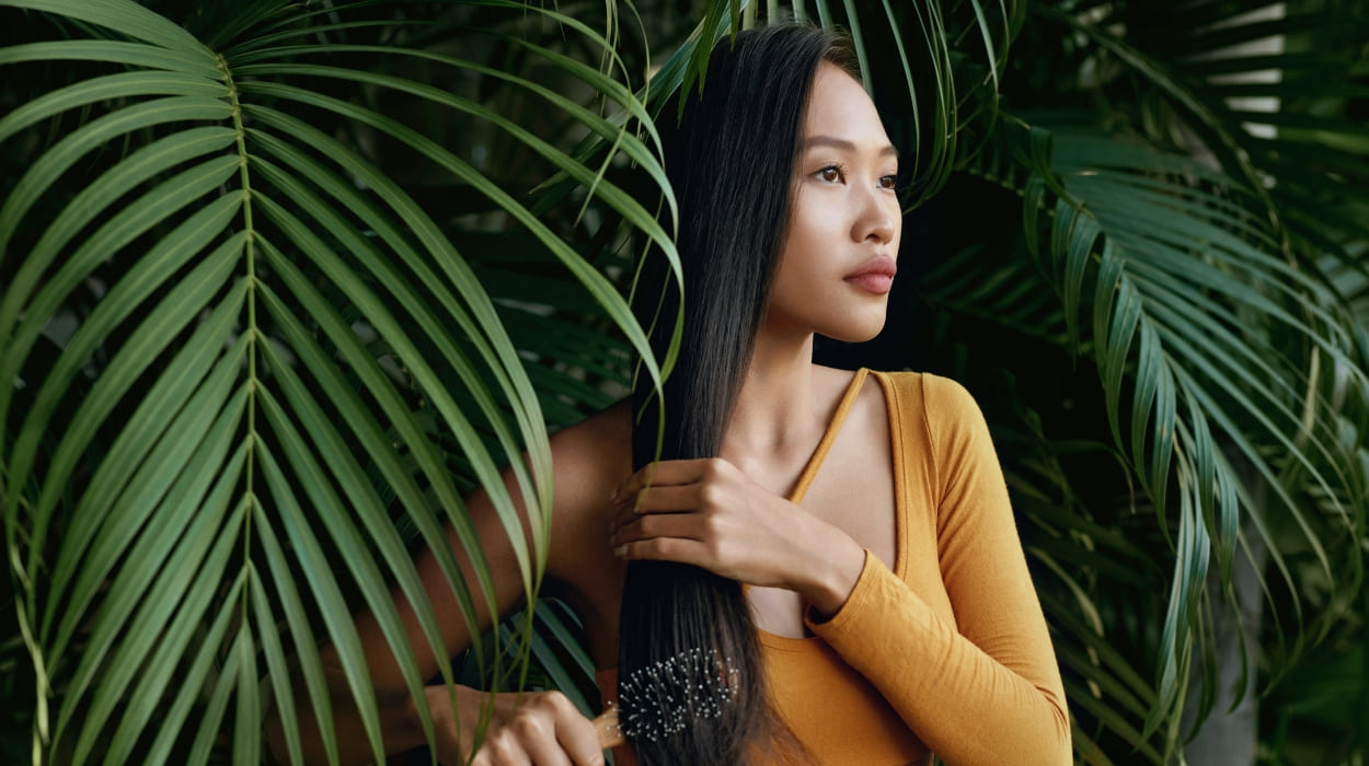 A woman brushes her long dark hair surrounded by palm trees